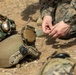 Military personnel in digital camouflage uniforms are gathered on arid ground, handling small components or wire with their hands. Multiple tactical helmets with communication headsets and rail-mounted accessories are visible on the ground. The scene appears to depict a training or preparation activity in a field environment.