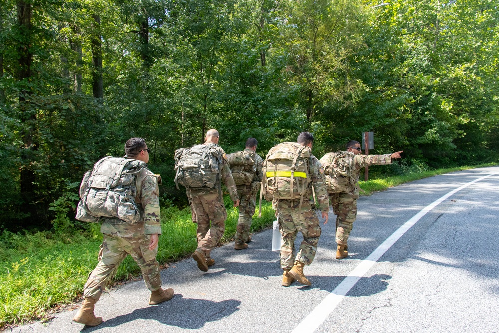 A group of five military personnel in camouflage uniforms are conducting a road march along a paved road bordered by dense green forest. They are carrying large rucksacks on their backs, and one soldier at the front right is pointing in a direction, likely providing navigation guidance. The scene appears to be a training exercise or ruck march event during daylight hours.