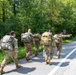 A group of five military personnel in camouflage uniforms are conducting a road march along a paved road bordered by dense green forest. They are carrying large rucksacks on their backs, and one soldier at the front right is pointing in a direction, likely providing navigation guidance. The scene appears to be a training exercise or ruck march event during daylight hours.