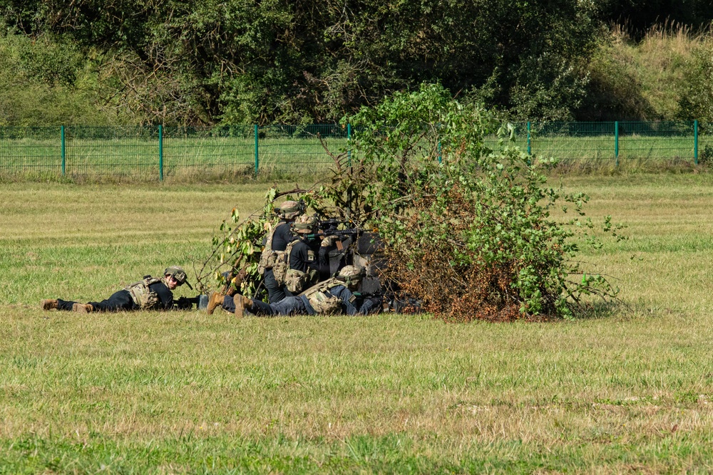 The image shows a group of military or tactical personnel in dark uniforms and combat gear taking covered positions behind a makeshift brush/foliage concealment in an open grassy field. At least one individual is prone on the ground to the left, while others are clustered behind the brush pile, with at least one appearing to aim a weapon. The setting appears to be a training exercise or demonstration, indicated by the fenced perimeter and well-maintained grounds.