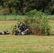 The image shows a group of military or tactical personnel in dark uniforms and combat gear taking covered positions behind a makeshift brush/foliage concealment in an open grassy field. At least one individual is prone on the ground to the left, while others are clustered behind the brush pile, with at least one appearing to aim a weapon. The setting appears to be a training exercise or demonstration, indicated by the fenced perimeter and well-maintained grounds.
