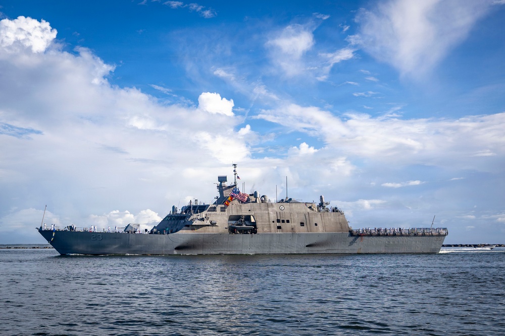 A U.S. Navy Littoral Combat Ship bearing hull number 23 is underway on open water under a partly cloudy sky. The vessel is flying the American flag and appears to have crew members visible on deck, suggesting a departure or arrival evolution. The ship displays the characteristic angular, low-profile design of the Freedom-class LCS.