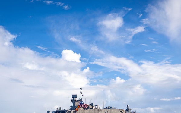 A U.S. Navy Littoral Combat Ship bearing hull number 23 is underway on open water under a partly cloudy sky. The vessel is flying the American flag and appears to have crew members visible on deck, suggesting a departure or arrival evolution. The ship displays the characteristic angular, low-profile design of the Freedom-class LCS.