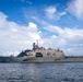 A U.S. Navy Littoral Combat Ship bearing hull number 23 is underway on open water under a partly cloudy sky. The vessel is flying the American flag and appears to have crew members visible on deck, suggesting a departure or arrival evolution. The ship displays the characteristic angular, low-profile design of the Freedom-class LCS.