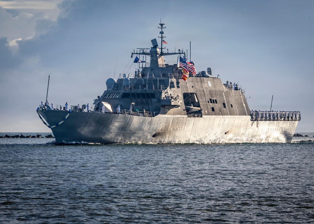 A U.S. Navy Littoral Combat Ship (LCS) is underway at sea, displaying the American flag and an additional colored flag (appearing yellow/gold and red) from its mast. The vessel features the distinctive angular, low-profile stealth hull design characteristic of the Freedom-class or Independence-class LCS. Crew members in white uniforms are visible along the deck rails.