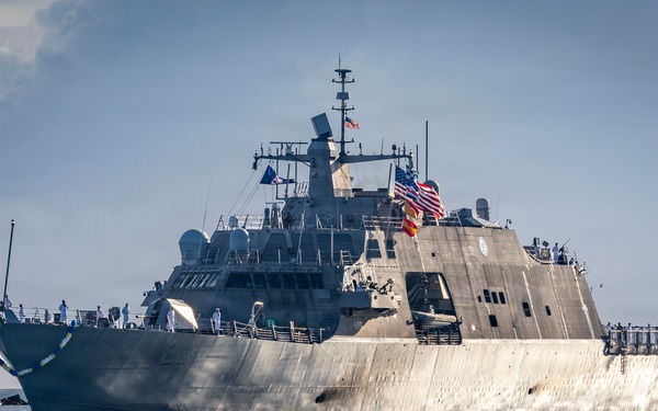 A U.S. Navy Littoral Combat Ship (LCS) is underway at sea, displaying the American flag and an additional colored flag (appearing yellow/gold and red) from its mast. The vessel features the distinctive angular, low-profile stealth hull design characteristic of the Freedom-class or Independence-class LCS. Crew members in white uniforms are visible along the deck rails.