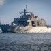 A U.S. Navy Littoral Combat Ship (LCS) is underway at sea, displaying the American flag and an additional colored flag (appearing yellow/gold and red) from its mast. The vessel features the distinctive angular, low-profile stealth hull design characteristic of the Freedom-class or Independence-class LCS. Crew members in white uniforms are visible along the deck rails.