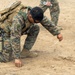 A U.S. Marine in full combat gear is kneeling on dry, sandy ground and appears to be examining or probing the soil with a small stick or probe. Additional Marines in matching MARPAT woodland/desert camouflage uniforms are visible in the background. The scene appears to be a field training exercise, possibly involving IED detection or ground inspection techniques.