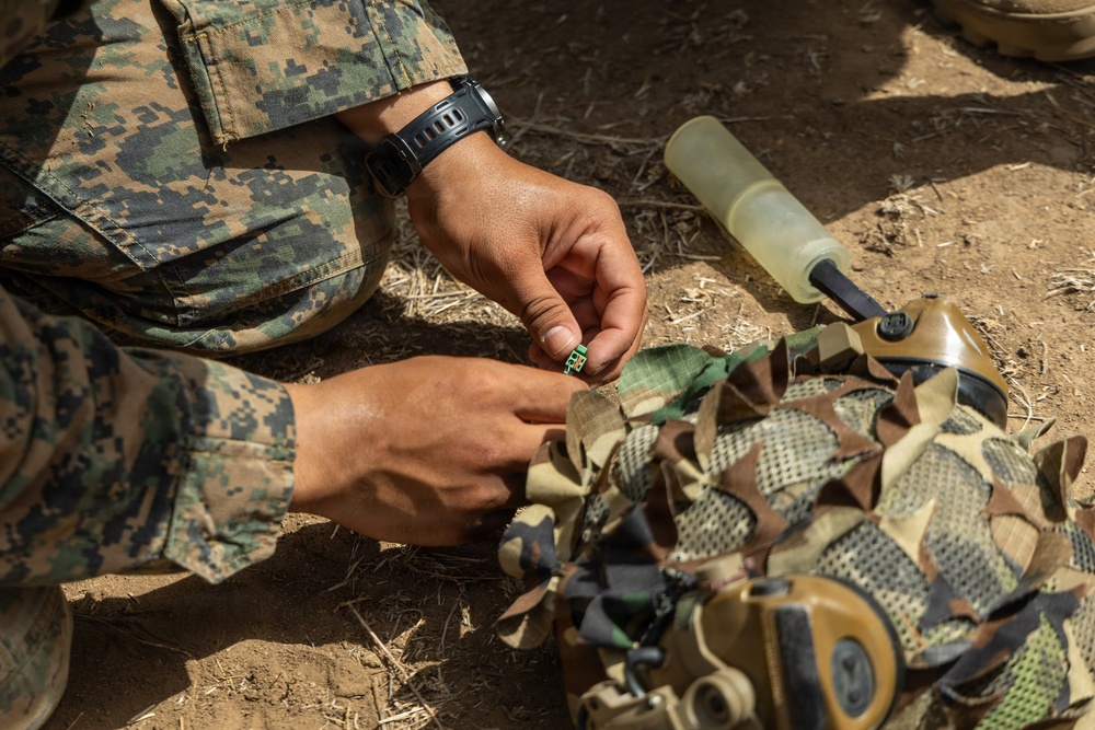 A military service member in MARPAT (Marine pattern) digital camouflage uniform is working with equipment on the ground. The individual appears to be assembling or attaching a small component to a camouflage-covered helmet or gear. A translucent cylindrical container is visible nearby on the dirt ground.