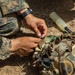 A military service member in MARPAT (Marine pattern) digital camouflage uniform is working with equipment on the ground. The individual appears to be assembling or attaching a small component to a camouflage-covered helmet or gear. A translucent cylindrical container is visible nearby on the dirt ground.