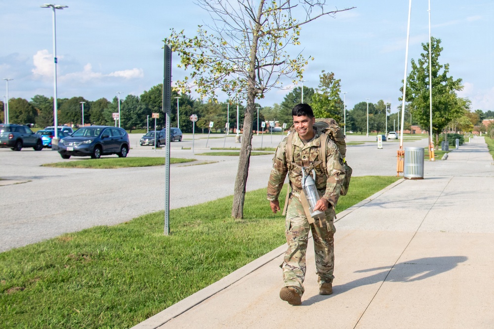 A smiling soldier in U.S. Army Operational Camouflage Pattern (OCP) uniform walks along a sidewalk on what appears to be a military installation. He is carrying a large rucksack on his back and holding a water bottle, suggesting he is completing or participating in a ruck march. The setting includes a large parking lot, lamp posts, trees, and well-maintained grounds consistent with a military base.