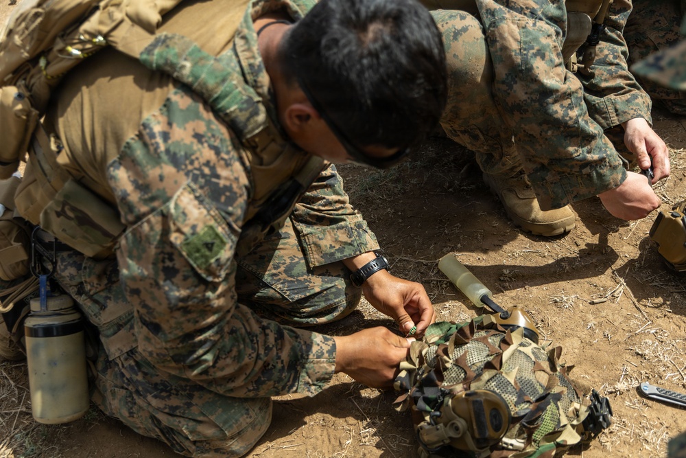 Two military personnel in MARPAT (Marine pattern) camouflage uniforms are kneeling on dry, sandy ground working with equipment. The foreground soldier appears to be assembling or adjusting gear, including what appears to be a camouflage helmet or netting with an attached tube or hose. The setting suggests a field training exercise or operational preparation.