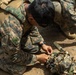 Two military personnel in MARPAT (Marine pattern) camouflage uniforms are kneeling on dry, sandy ground working with equipment. The foreground soldier appears to be assembling or adjusting gear, including what appears to be a camouflage helmet or netting with an attached tube or hose. The setting suggests a field training exercise or operational preparation.