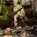 Two military personnel in camouflage uniforms and tactical gear are operating electronic equipment in a field setting with vegetation cover. They appear to be working with a control device and cabling connected to a black equipment case. The setting suggests a field training exercise or operational deployment.