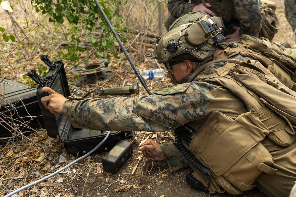 A military personnel wearing MARPAT digital camouflage and tactical gear is operating what appears to be a ruggedized laptop or ground control station in a field environment. The soldier is prone or crouching among leaf litter and sparse vegetation, manipulating cables and controls connected to the device. The setting suggests a field training exercise or operational deployment involving electronic or communications equipment.
