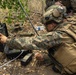 A military personnel wearing MARPAT digital camouflage and tactical gear is operating what appears to be a ruggedized laptop or ground control station in a field environment. The soldier is prone or crouching among leaf litter and sparse vegetation, manipulating cables and controls connected to the device. The setting suggests a field training exercise or operational deployment involving electronic or communications equipment.