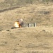 An explosion is occurring on a dry, grassy hillside near what appears to be a vehicle and a large metal shipping container or structure. The scene shows an active fireball with smoke rising from the point of detonation. Debris is scattered across the surrounding terrain, suggesting this may be a controlled demolition or weapons test on a range.