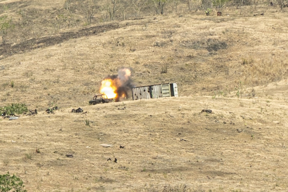An explosion is occurring on a dry, grassy hillside near what appears to be a vehicle and a large metal shipping container or structure. The scene shows an active fireball with smoke rising from the point of detonation. Debris is scattered across the surrounding terrain, suggesting this may be a controlled demolition or weapons test on a range.