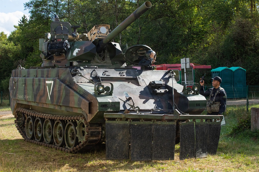 A tracked military armored fighting vehicle in multi-tone camouflage paint is stationary on grass, with at least two personnel visible — one in the turret and one standing beside the vehicle. The vehicle appears to be equipped with a large-caliber gun and side skirt armor panels. The setting appears to be a military exercise, demonstration, or display event, with civilian infrastructure visible in the background.