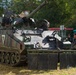 A tracked military armored fighting vehicle in multi-tone camouflage paint is stationary on grass, with at least two personnel visible — one in the turret and one standing beside the vehicle. The vehicle appears to be equipped with a large-caliber gun and side skirt armor panels. The setting appears to be a military exercise, demonstration, or display event, with civilian infrastructure visible in the background.