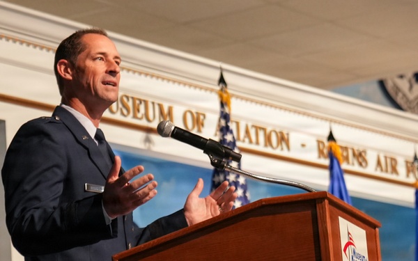 A uniformed U.S. Air Force officer is delivering a speech at a wooden podium bearing the 'Museum of Aviation' logo. The event is taking place at the Museum of Aviation at Robins Air Force Base, as indicated by the text visible on the wall behind the speaker. Multiple flags, including U.S. and Air Force flags, are displayed in the background.