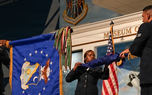 Three uniformed military personnel are participating in a formal flag ceremony, unfolding or presenting a large blue ceremonial flag with gold fringe and an eagle emblem. The event appears to be taking place inside or in front of a museum facility, with a U.S. flag and military insignia visible in the background. Multiple colored streamers or battle streamers are attached to the flag's staff.