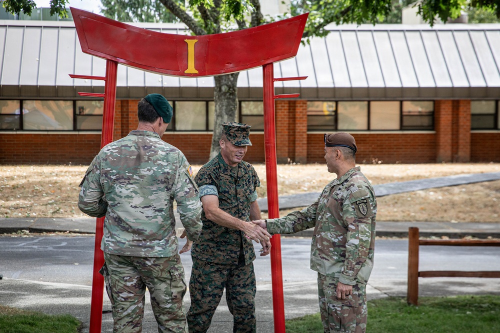 Three military personnel in uniform are gathered beneath a red torii-style gate structure featuring a yellow Roman numeral 'I'. The central figure, wearing Marine Corps MARPAT camouflage and cover, is shaking hands with a soldier on the right wearing OCP camouflage and a tan beret. A third soldier wearing OCP camouflage and a green beret stands to the left.