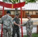 Three military personnel in uniform are gathered beneath a red torii-style gate structure featuring a yellow Roman numeral 'I'. The central figure, wearing Marine Corps MARPAT camouflage and cover, is shaking hands with a soldier on the right wearing OCP camouflage and a tan beret. A third soldier wearing OCP camouflage and a green beret stands to the left.