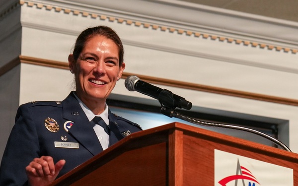 A female military officer in a dark blue dress uniform is speaking at a podium during an event at the Museum of Aviation. She is smiling and appears engaged with her audience. The setting suggests a formal ceremony or official speaking engagement.