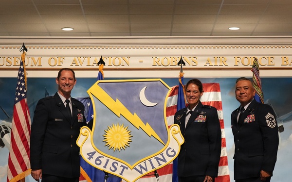 Three U.S. Air Force personnel pose on a stage at the Museum of Aviation at Robins Air Force Base during what appears to be an official military ceremony. They are standing behind a display shield bearing the insignia and name '461st Wing'. The setting includes American flags and a unit guidon flag.