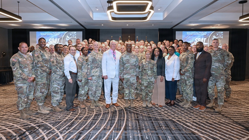 A large group photo taken in a hotel ballroom or conference center featuring predominantly U.S. Army personnel in OCP (Operational Camouflage Pattern) uniforms alongside several civilians in business attire. Two presentation screens in the background display 'SDAT' branding with text reading 'Senior Contracting Officials and Directors Acquisition Training.' The group appears to be assembled for a formal military acquisition training event.