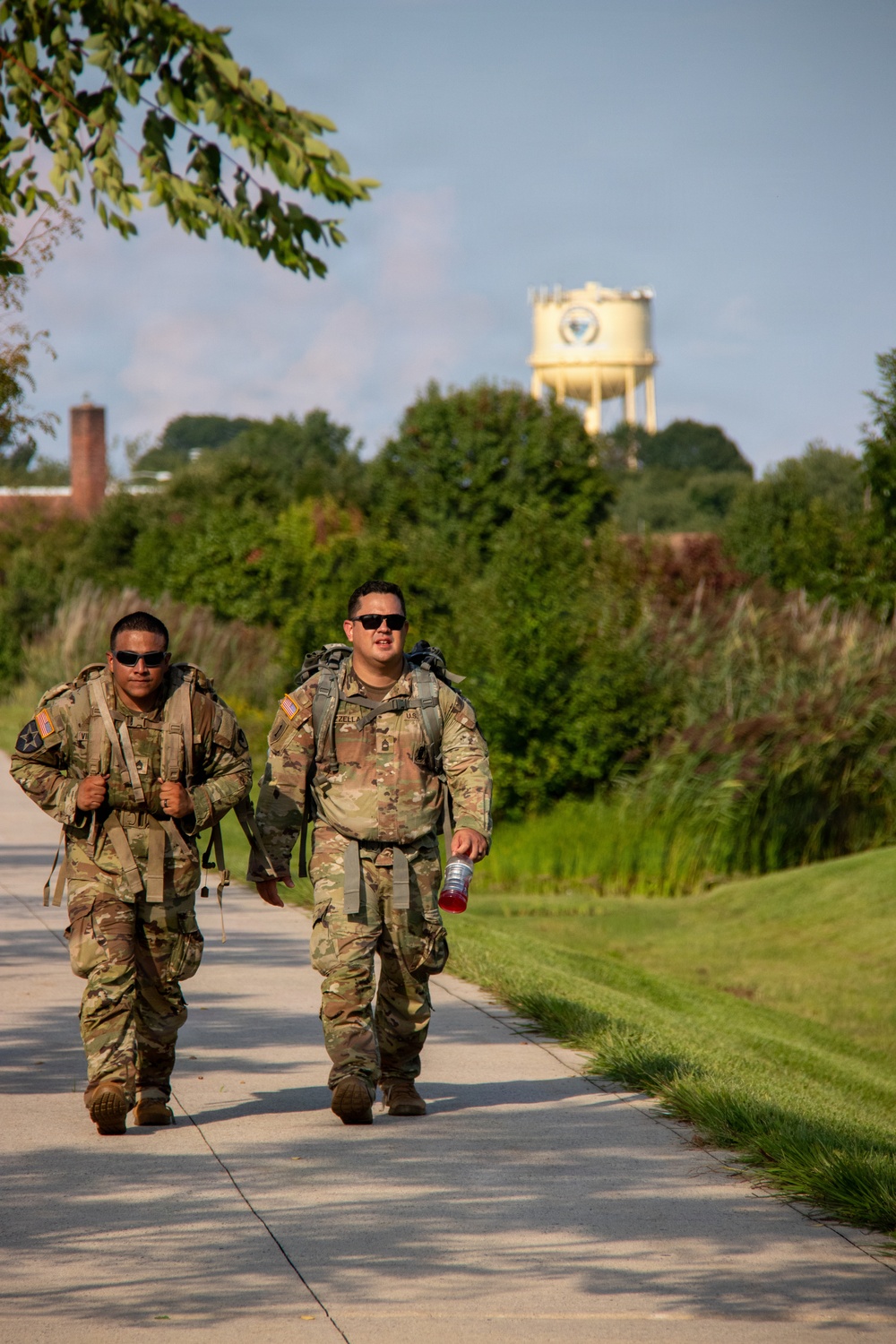 Two U.S. Army soldiers in OCP (Operational Camouflage Pattern) uniforms are conducting a ruck march along a paved pathway on what appears to be a military installation. Both soldiers are wearing full rucksacks and sunglasses, with one carrying a red water bottle. A military water tower with an insignia is visible in the background.