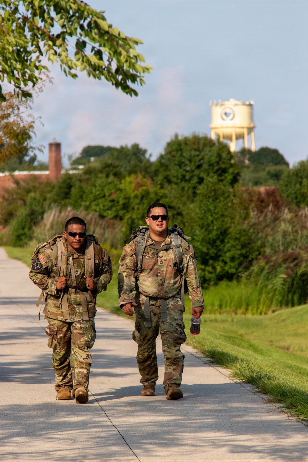 Two U.S. Army soldiers in Operational Camouflage Pattern (OCP) uniforms are walking along a paved path while carrying rucksacks, likely conducting a ruck march. Both wear sunglasses and have American flag patches on their right shoulders. The background features green vegetation, trees, and a distinctive water tower.