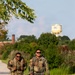 Two U.S. Army soldiers in Operational Camouflage Pattern (OCP) uniforms are walking along a paved path while carrying rucksacks, likely conducting a ruck march. Both wear sunglasses and have American flag patches on their right shoulders. The background features green vegetation, trees, and a distinctive water tower.