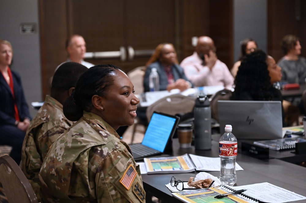 A U.S. Army soldier in OCP (Operational Camouflage Pattern) uniform is seated at a table during what appears to be a professional meeting or training session. She is turned slightly and appears to be speaking or engaged in discussion. The room contains multiple civilian and military participants seated at tables with laptops, notebooks, and water bottles.