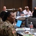 A U.S. Army soldier in OCP (Operational Camouflage Pattern) uniform is seated at a table during what appears to be a professional meeting or training session. She is turned slightly and appears to be speaking or engaged in discussion. The room contains multiple civilian and military participants seated at tables with laptops, notebooks, and water bottles.