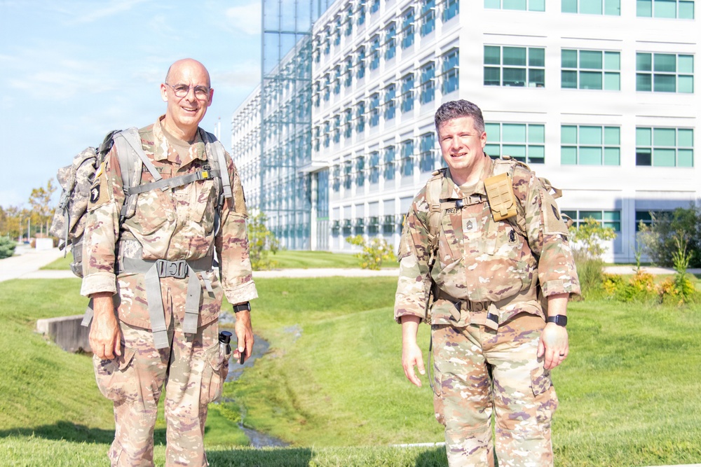 Two military personnel in U.S. Army OCP (Operational Camouflage Pattern) uniforms are posing outdoors in front of a large modern glass-and-steel building. Both are wearing rucksacks and tactical gear, suggesting they are participating in a ruck march or physical training event. The setting appears to be a military installation or government campus on a clear, sunny day.