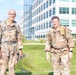 Two military personnel in U.S. Army OCP (Operational Camouflage Pattern) uniforms are posing outdoors in front of a large modern glass-and-steel building. Both are wearing rucksacks and tactical gear, suggesting they are participating in a ruck march or physical training event. The setting appears to be a military installation or government campus on a clear, sunny day.