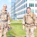 Two U.S. Army soldiers in OCP (Operational Camouflage Pattern) uniforms are walking outdoors near a modern glass office building. Both are wearing rucksacks and appear to be in good spirits, smiling at the camera. The setting suggests a military installation or government campus.