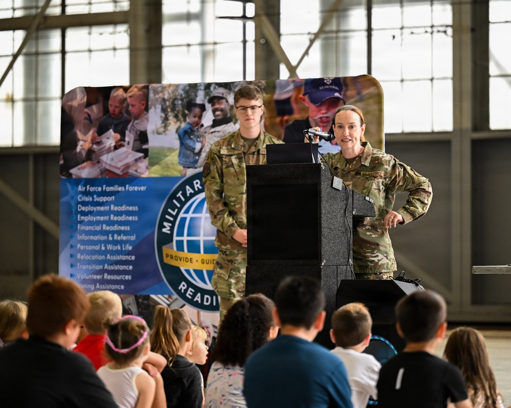 A female U.S. Air Force service member is speaking at a podium to a group of children seated on the floor. The event appears to be a Military Readiness program presentation, with a branded banner visible behind the speakers. A second service member stands behind the podium as the presenter addresses the young audience.