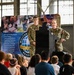 A female U.S. Air Force service member is speaking at a podium to a group of children seated on the floor. The event appears to be a Military Readiness program presentation, with a branded banner visible behind the speakers. A second service member stands behind the podium as the presenter addresses the young audience.