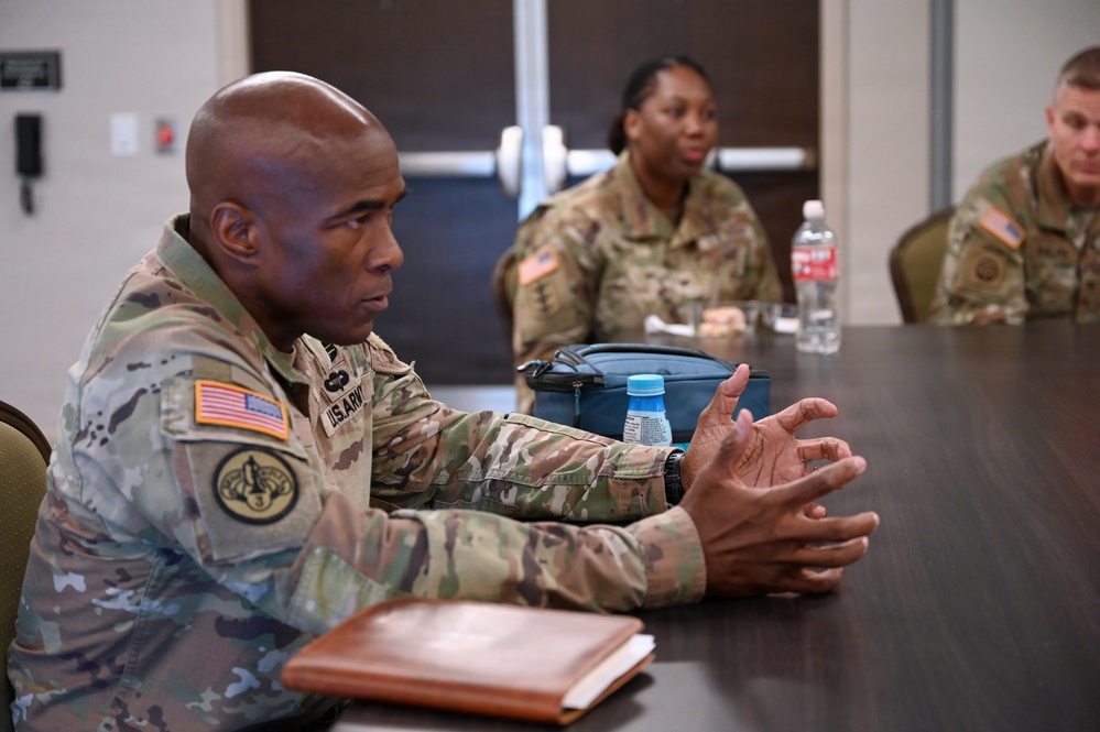 Three U.S. Army soldiers in Operational Camouflage Pattern (OCP) uniforms are seated at a conference table during what appears to be a meeting or briefing. The soldier in the foreground is actively speaking and gesturing with his hands, while two other soldiers are visible in the background. The setting appears to be an indoor meeting room or conference facility.