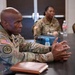 Three U.S. Army soldiers in Operational Camouflage Pattern (OCP) uniforms are seated at a conference table during what appears to be a meeting or briefing. The soldier in the foreground is actively speaking and gesturing with his hands, while two other soldiers are visible in the background. The setting appears to be an indoor meeting room or conference facility.