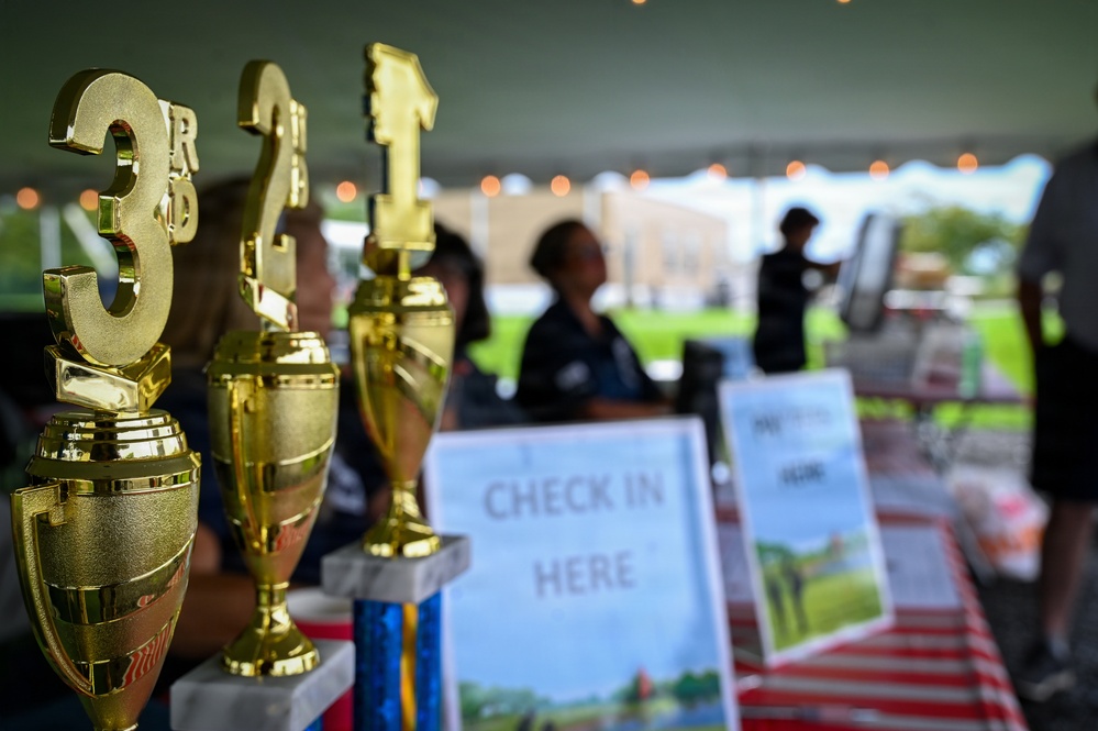 The image shows a registration or check-in table at an outdoor event, likely a competition or tournament, set up under a tent. Three gold trophies marked 1st, 2nd, and 3rd place are prominently displayed in the foreground. A 'CHECK IN HERE' sign is visible on the table, with blurred figures of attendees and a building visible in the background.