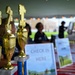 The image shows a registration or check-in table at an outdoor event, likely a competition or tournament, set up under a tent. Three gold trophies marked 1st, 2nd, and 3rd place are prominently displayed in the foreground. A 'CHECK IN HERE' sign is visible on the table, with blurred figures of attendees and a building visible in the background.
