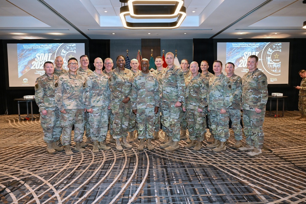 A group of approximately 15-16 U.S. Army personnel in MultiCam/OCP camouflage uniforms pose for a formal group photo at what appears to be a military training event. The event is identified by projected screens as 'SDAT 2024' (Senior Contracting Officials and Directors Acquisition Training). The setting is a hotel or conference center ballroom with U.S. and military flags displayed in the background.