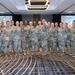 A group of approximately 15-16 U.S. Army personnel in MultiCam/OCP camouflage uniforms pose for a formal group photo at what appears to be a military training event. The event is identified by projected screens as 'SDAT 2024' (Senior Contracting Officials and Directors Acquisition Training). The setting is a hotel or conference center ballroom with U.S. and military flags displayed in the background.