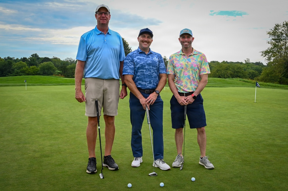 Three adult men are posing together on a golf course putting green, each holding a putter. They are dressed in casual golf attire and appear to be participating in a recreational or charity golf outing. The background shows a well-maintained fairway, trees, and a partly cloudy sky.