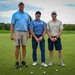 Three adult men are posing together on a golf course putting green, each holding a putter. They are dressed in casual golf attire and appear to be participating in a recreational or charity golf outing. The background shows a well-maintained fairway, trees, and a partly cloudy sky.