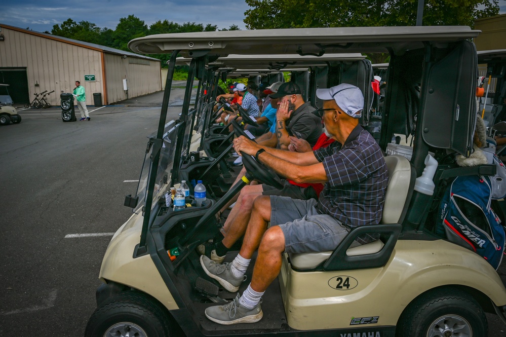 A line of Yamaha golf carts, numbered and staged in a parking area, are loaded with golfers preparing for a shotgun-start tournament or group outing. The foreground cart (#24) seats two men wearing caps and casual golf attire, with water bottles and scorecards visible. A person in a green shirt walks in the background near a storage building.